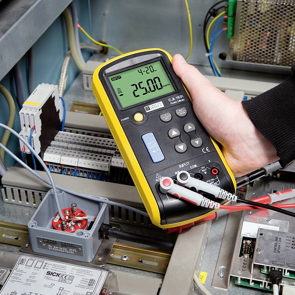 Dłoń trzyma multimetr, który wskazuje 25,0 mA, będąc podłączonym do elektrycznej tablicy rozdzielczej z wieloma przewodami i elementami.