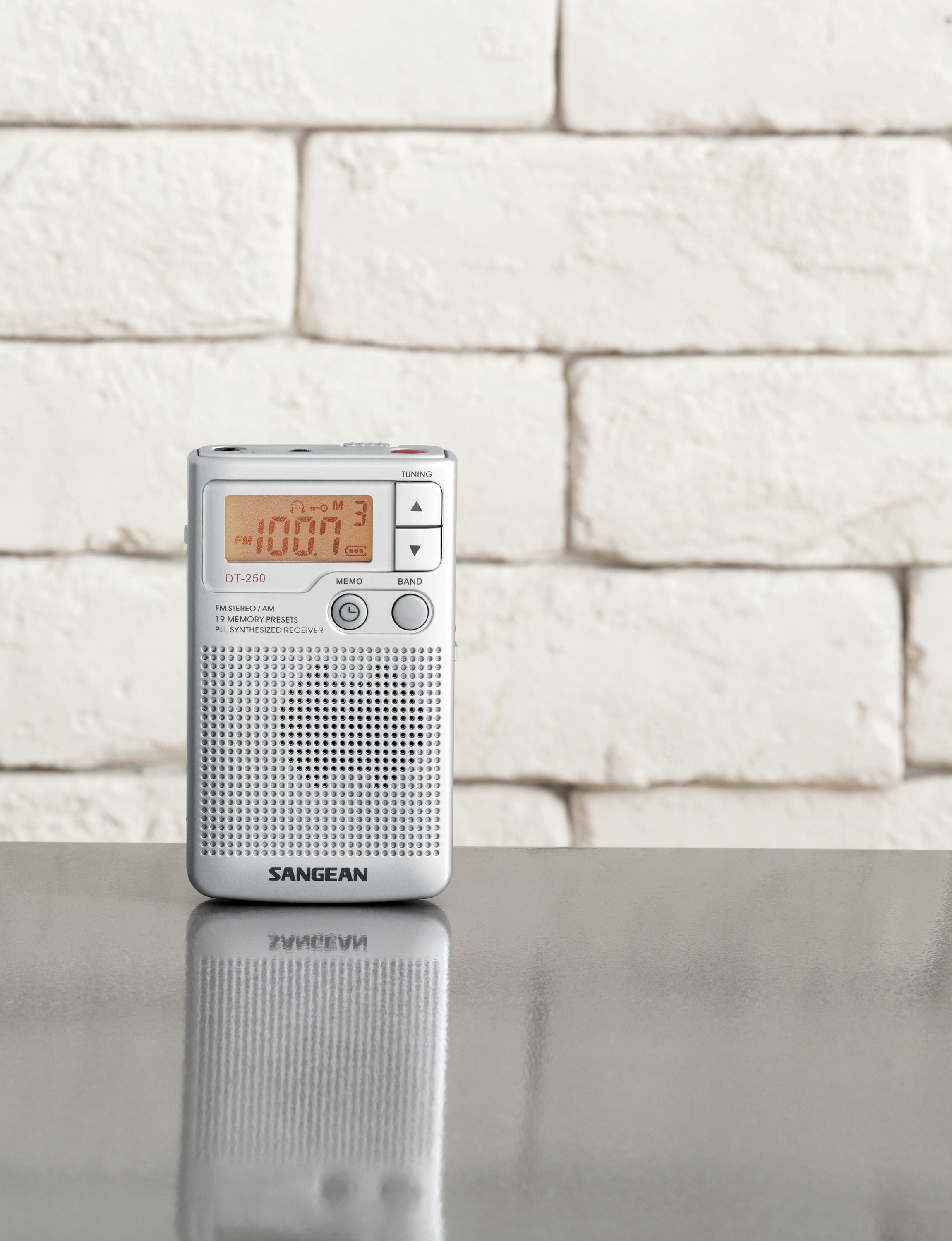 Mały przenośny radio stoi na stole przed białą ścianą z cegły. Na wyświetlaczu widać 100.7.