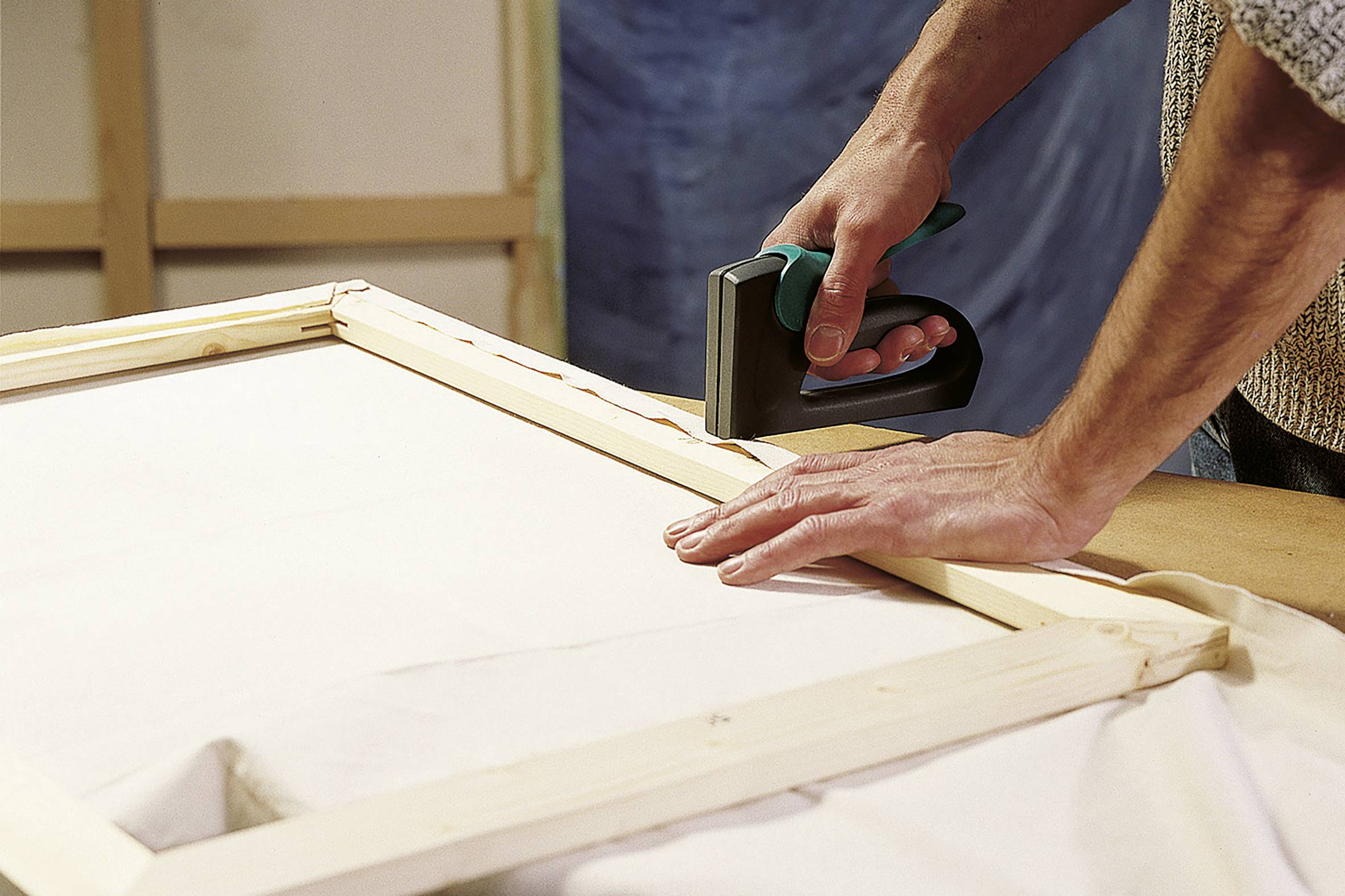 Osoba mocuje płótno do drewnianej ramy za pomocą zszywacza. Ręce trzymają ramę mocno. W tle widoczne jest środowisko pracy.