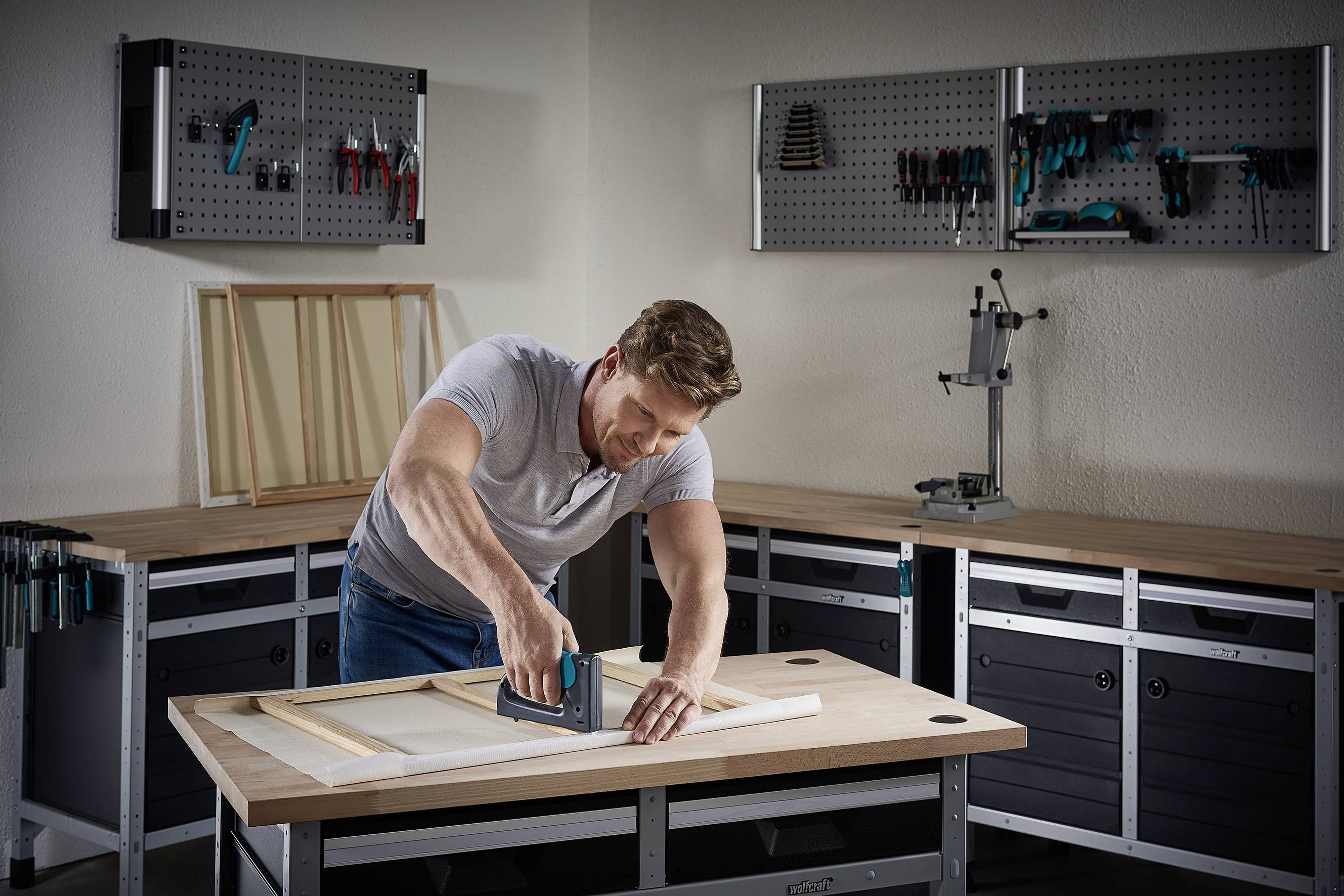 Mężczyzna używa zszywacza przy drewnianej ramie w warsztacie, na którego ścianach i stołach warsztatowych znajdują się różne narzędzia.