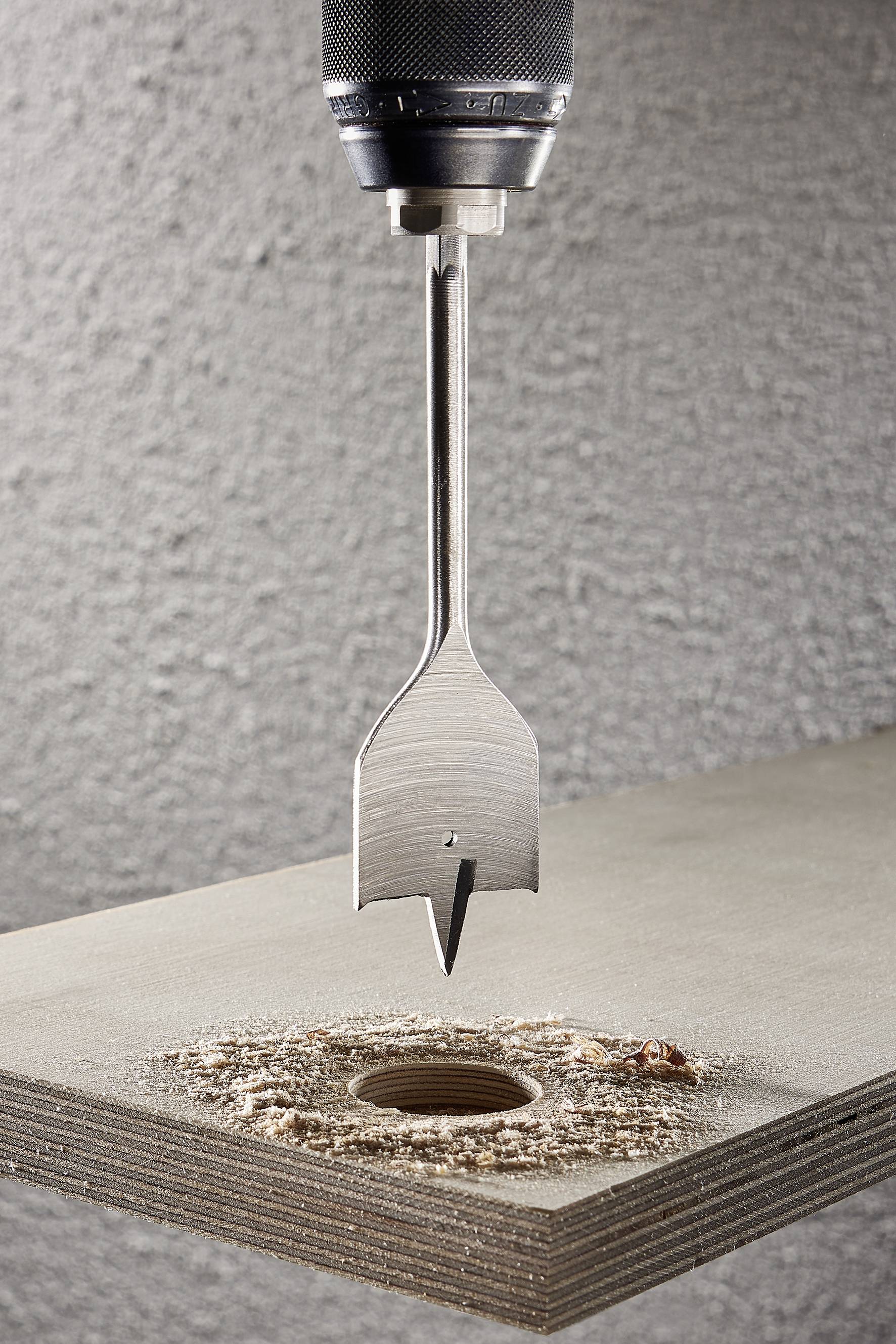 Wiertarka z płaskim świdrowym końcówką wierci otwór w desce. Wokół otworu widoczne są wióry.