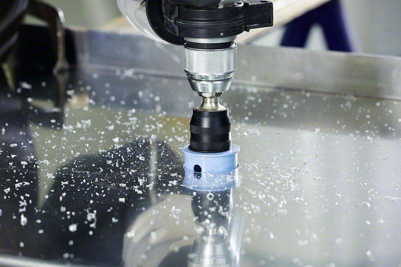 Elektryczna wiertarka z końcówką do wycinania otworów wierci dziurę w blasze metalowej. Metalowe odpryski są rozrzucone wokół miejsca wiercenia.