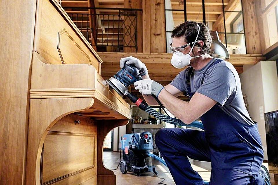 Mężczyzna w odzieży ochronnej szlifuje drewniany element mebla w warsztacie. Na tle widać narzędzia.