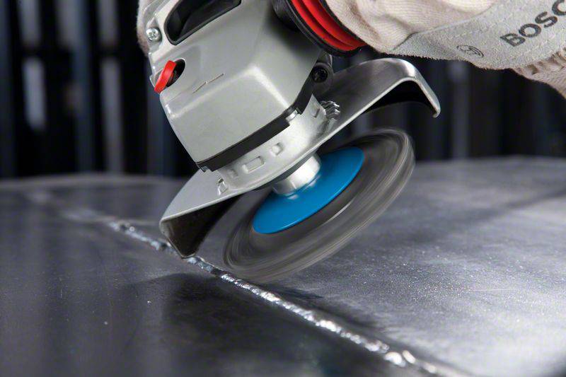 Szlifierka kątowa z niebieskim kołem szlifierskim wygładza spoinę spawalniczą na powierzchni metalowej. Ręka w rękawicy trzyma narzędzie.