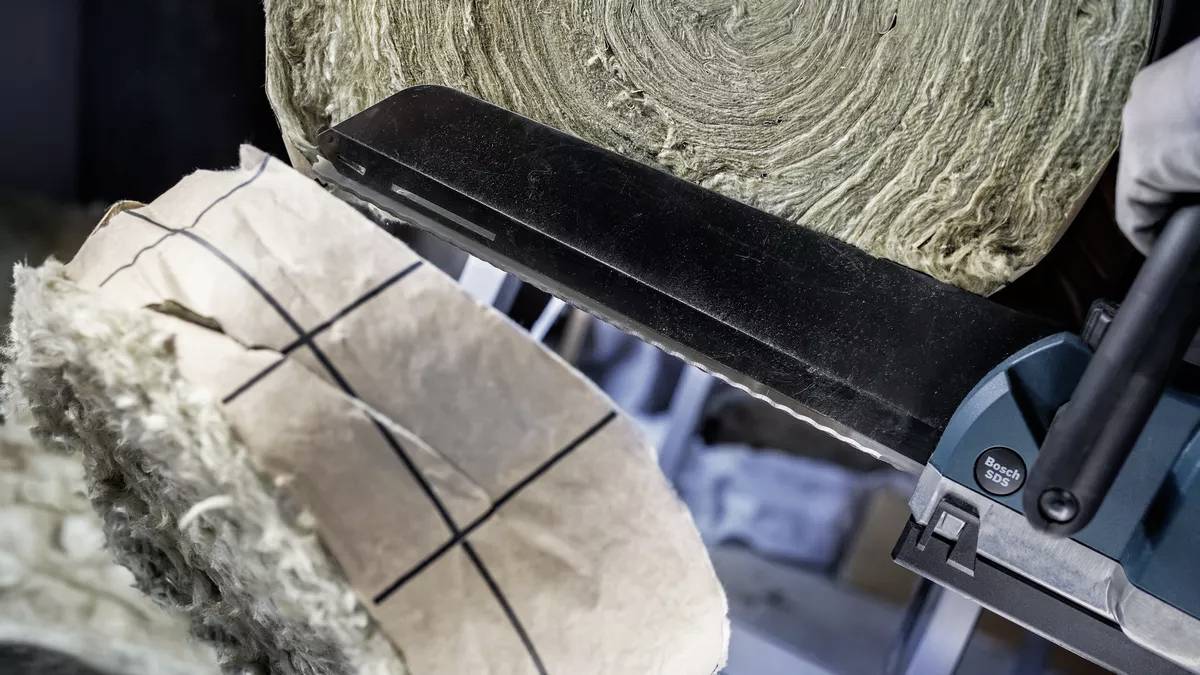 Osoba przecina elektryczną piłą rolkę materiału izolacyjnego. Cząsteczki pyłu unoszą się w powietrzu.