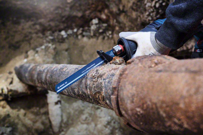 Dłoń trzymająca elektryczną piłę, która przecina skorodowaną i kruchą metalową rurę pod ziemią.