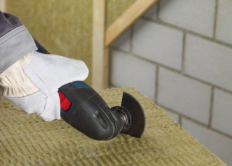 Rzemieślnik używa narzędzia oscylacyjnego do cięcia materiału izolacyjnego. Na tle widoczna jest ceglana ściana.