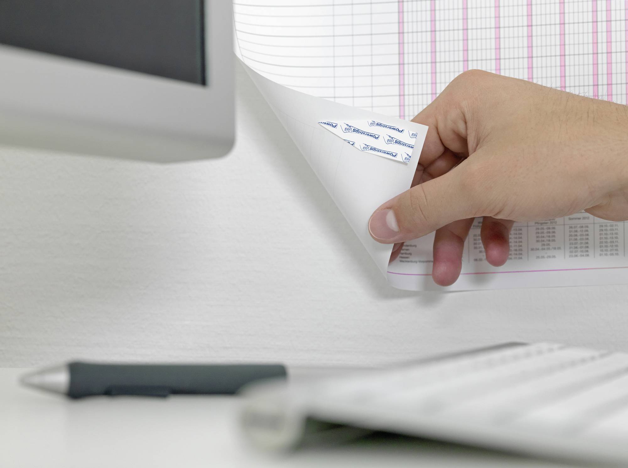 Dłoń przewraca stronę kalendarza, obok widoczny jest monitor, klawiatura i długopis.