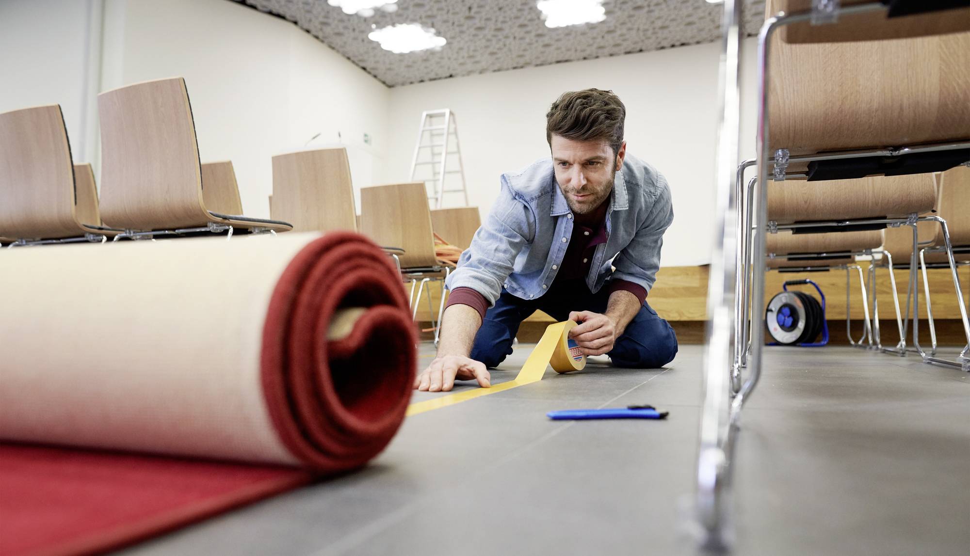 Mężczyzna kładzie wykładzinę dywanową w sali konferencyjnej. Klęczy na podłodze i używa taśmy klejącej. W tle widać krzesła.