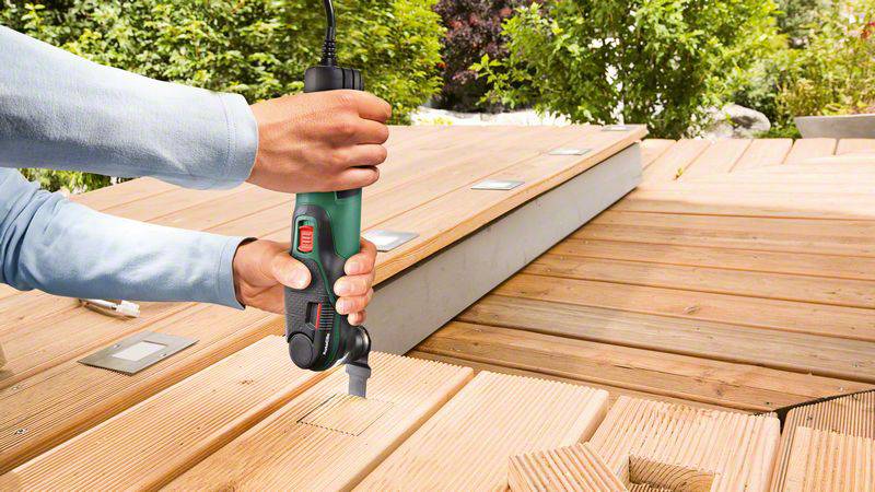 Jedna osoba używa elektrycznego wielofunkcyjnego narzędzia do wycinania prostokątnego otworu w drewnianym tarasie.