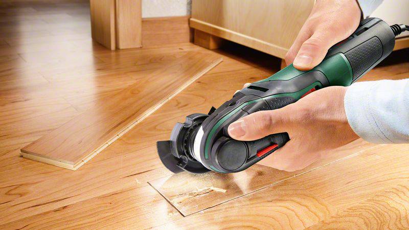 Osoba przycina elektrycznym narzędziem drewnianą płytę. Na podłodze leżą już wcześniej pocięte kawałki drewna.