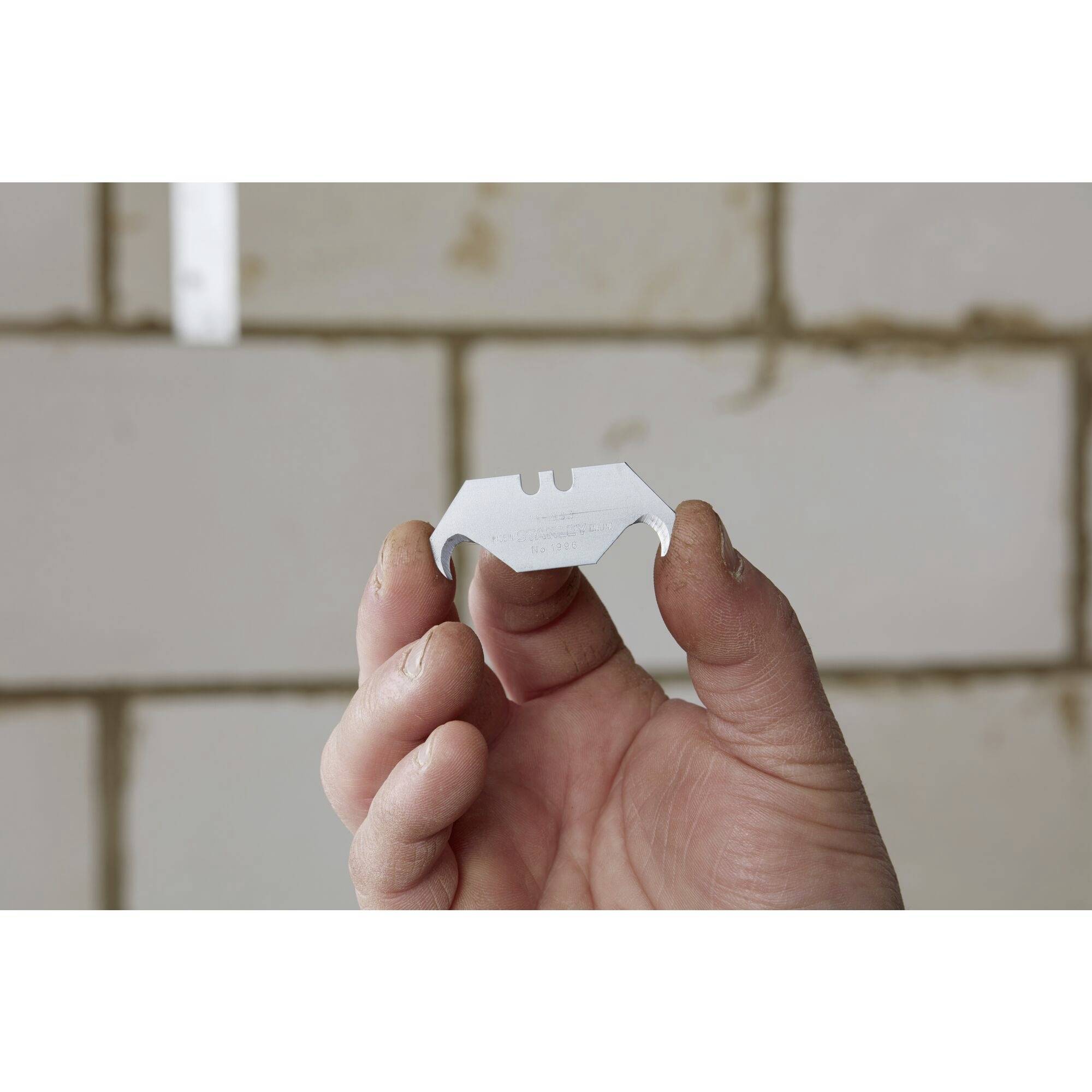 Dłoń trzyma ostrze żyletki przed rozmytym murem z cegły. Ostrze jest delikatnie przytrzymywane opuszkami palców.