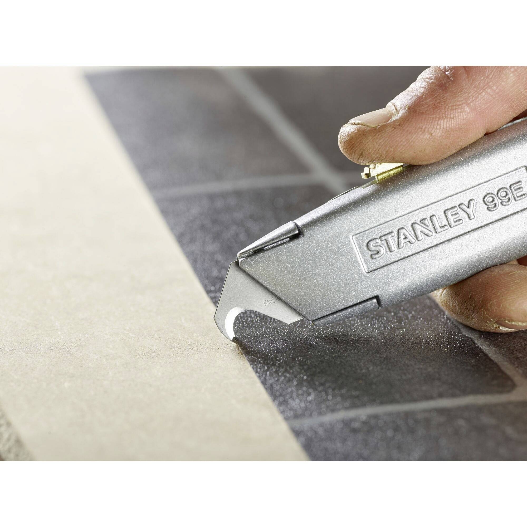 Ręka trzyma srebrny nóż uniwersalny Stanley-99E na szarej powierzchni, gotowy do cięcia.