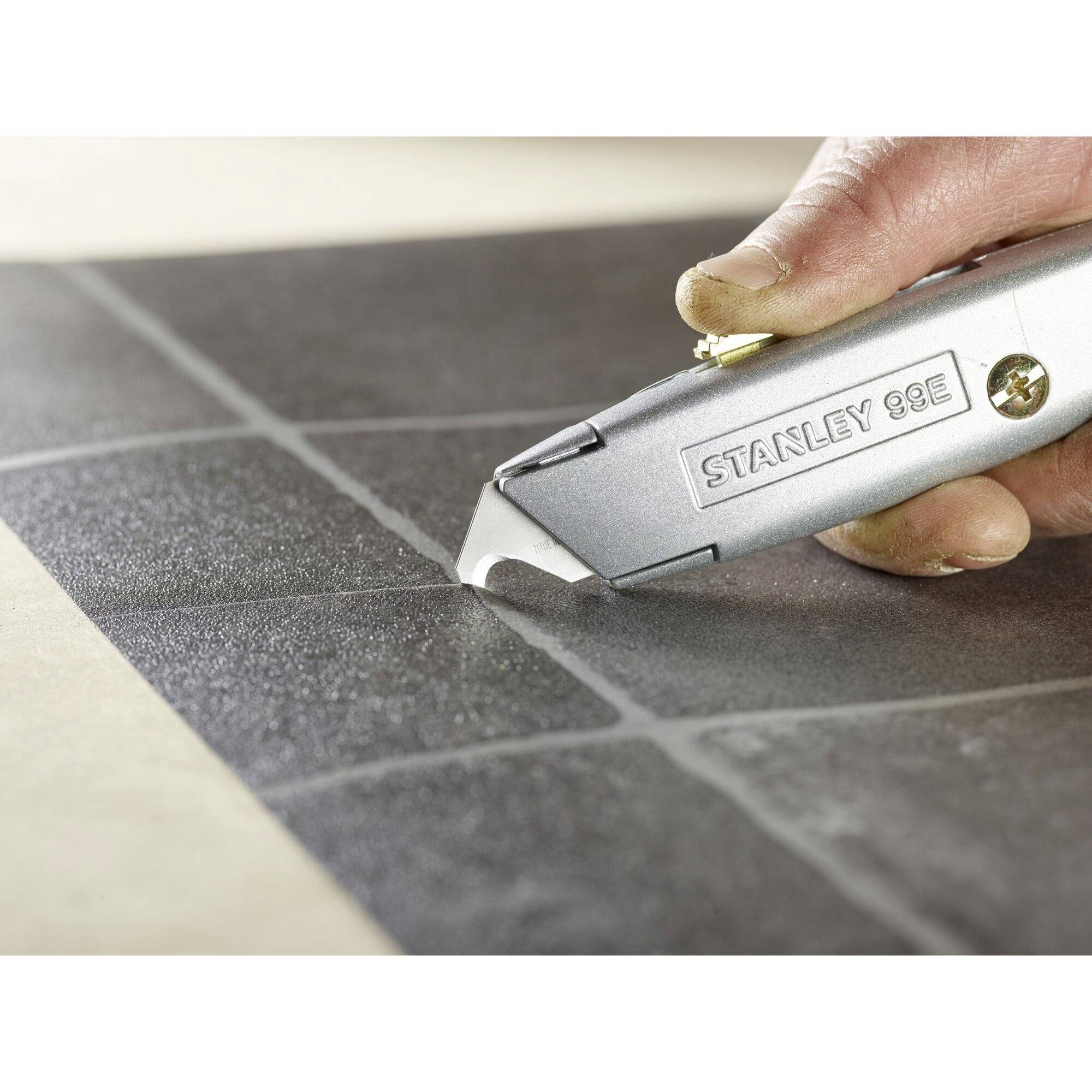 Ręka trzyma srebrny nóż uniwersalny z napisem 'Stanley 99E', precyzyjnie przecinający czarną powierzchnię płytki.