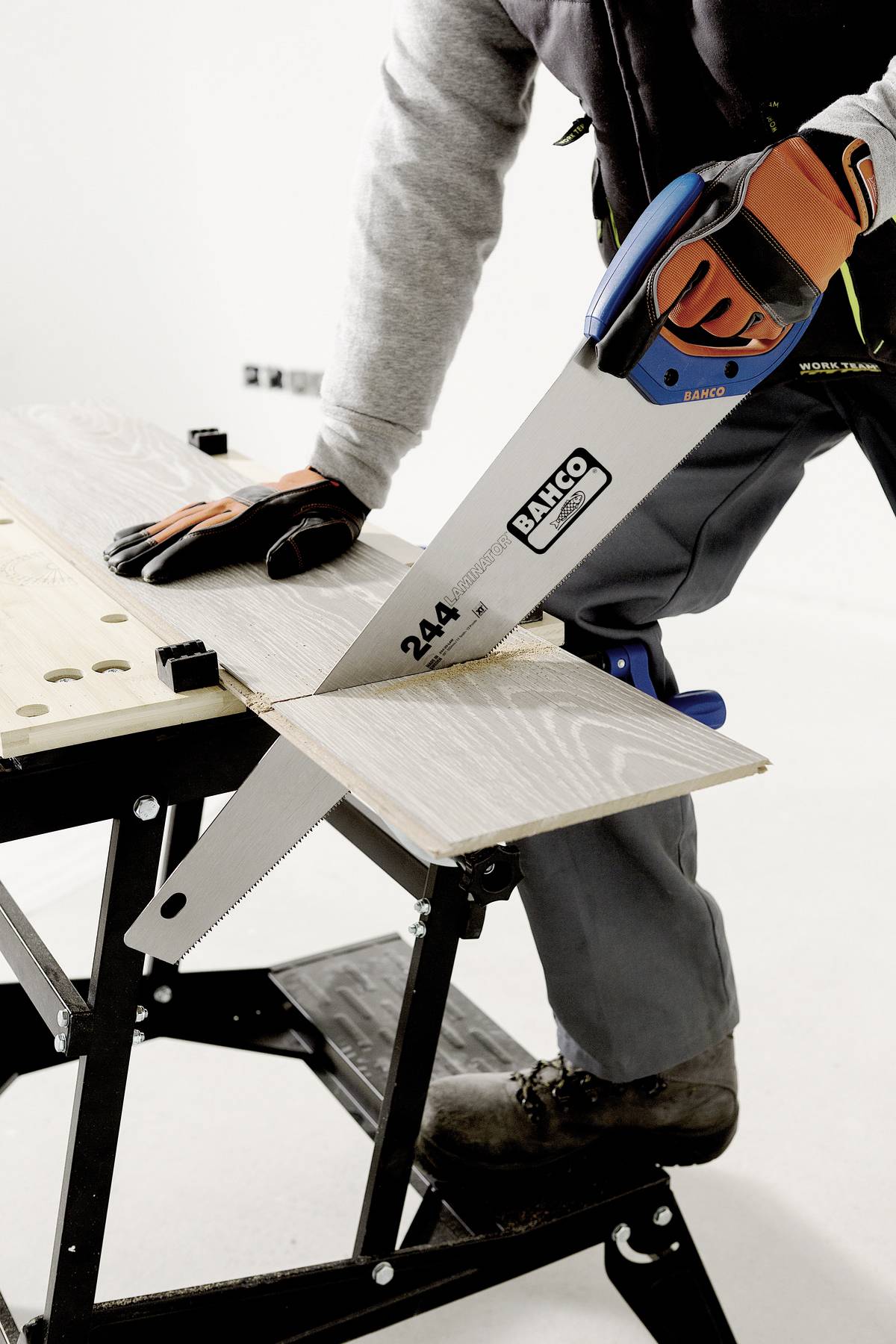 Osoba piłuje deskę drewnianą na koźle stolarskim za pomocą piły ręcznej. Osoba ma na sobie rękawice robocze i kurtkę roboczą.