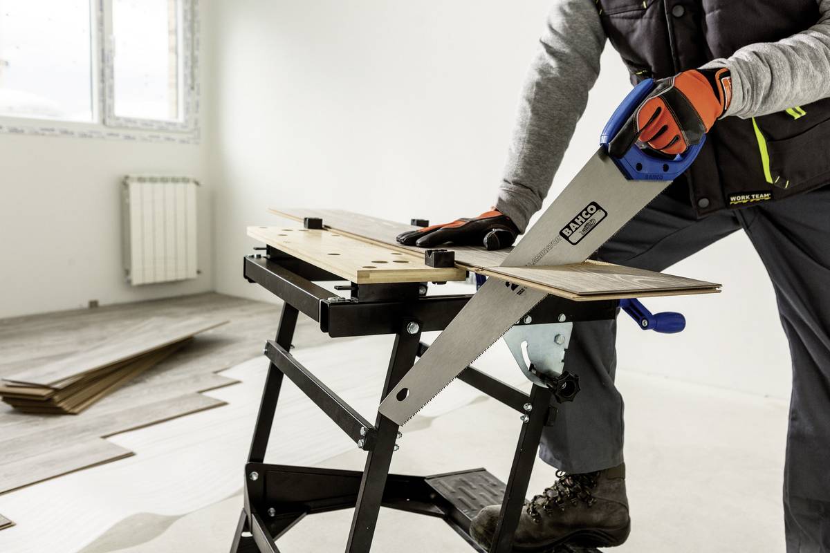 Osoba piłuje deskę na przenośnym stole roboczym w pustym pomieszczeniu. W tle leżą deski podłogowe.