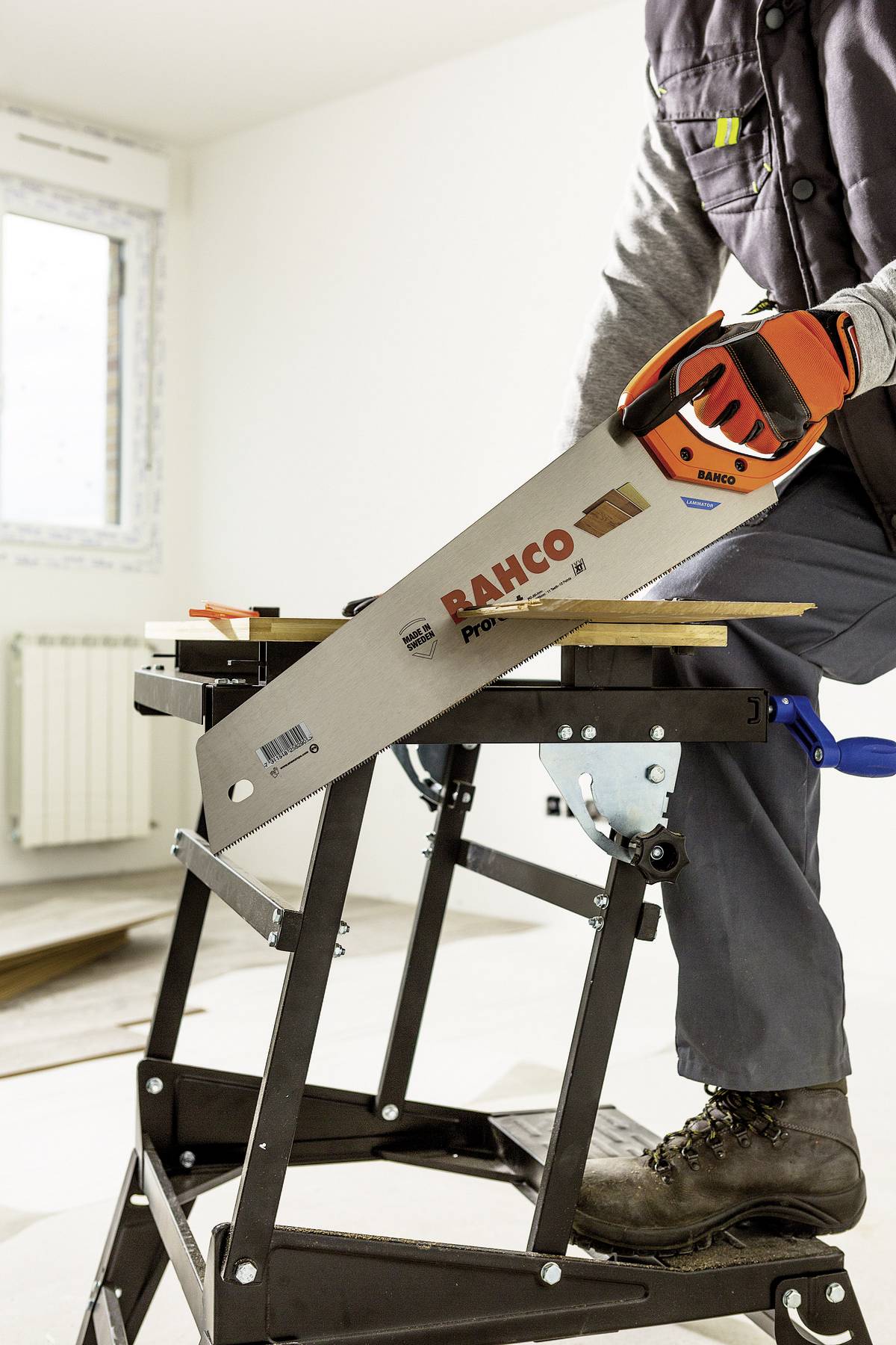 Osoba piłuje deską drewnianą ręczną piłą na składanym stole w jasnym pomieszczeniu. Na pile widnieje napis 'RAHCO'.