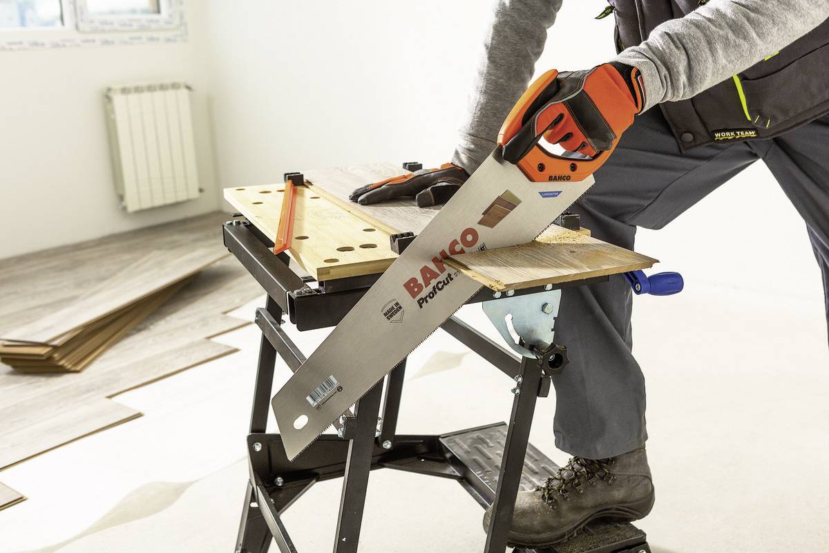 Osoba piłuje kawałek drewna ręczną piłą na stole warsztatowym. W tle widoczne są deski laminowane.