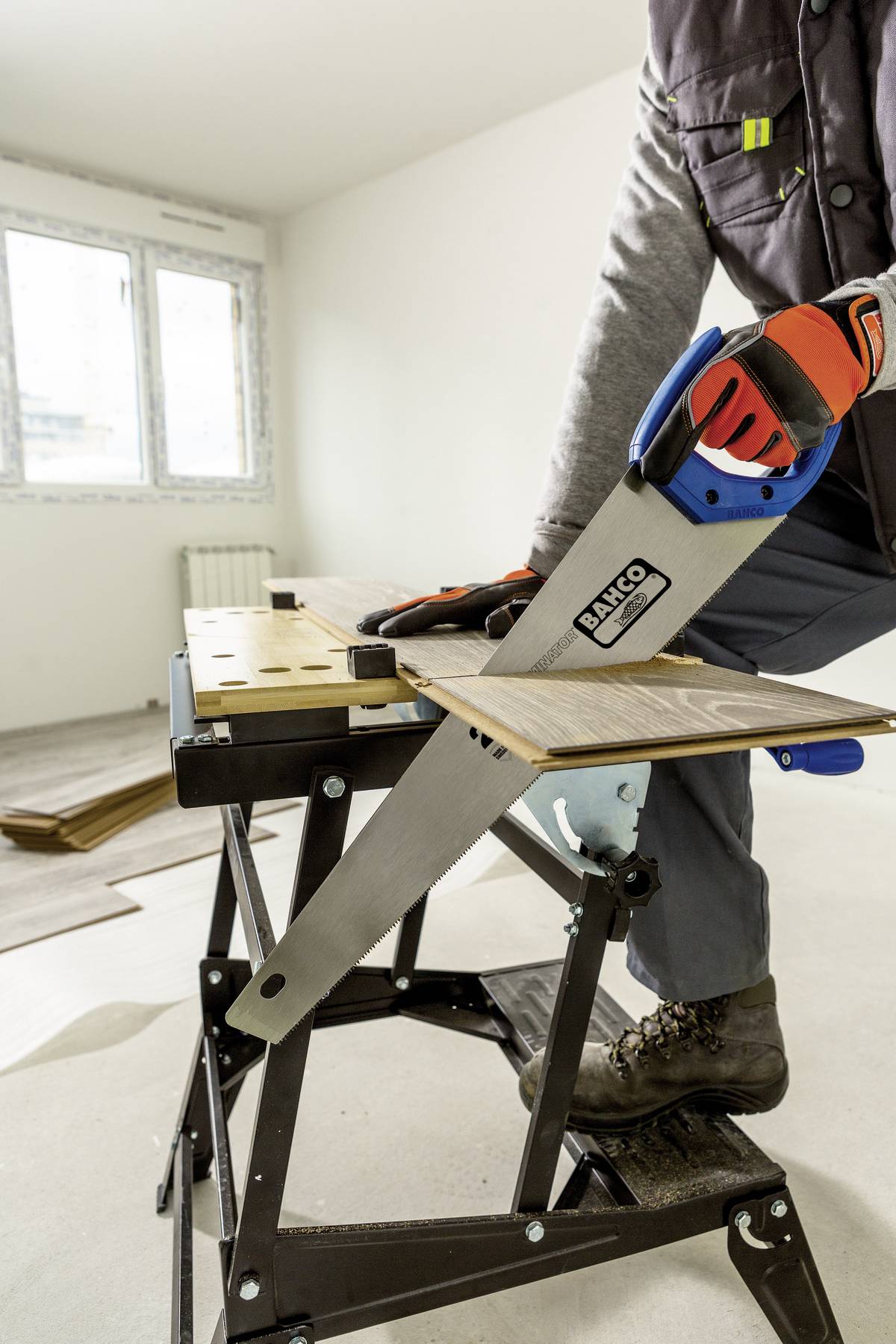 Osoba piłuje drewno ręczną piłą na stole warsztatowym w wyremontowanym pomieszczeniu. Ma na rękach rękawice robocze.