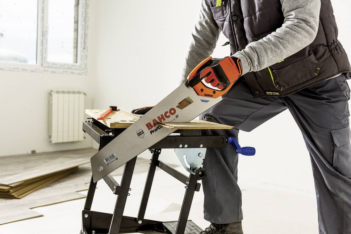 Osoba przecina deskę drewnianą piłą ręczną na jasnym tle, na stole warsztatowym. Trwają prace remontowe lub budowlane.