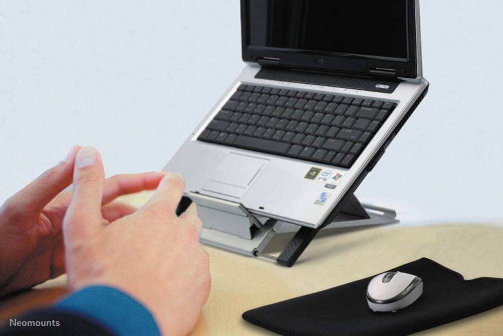 Laptop stoi na regulowanym stojaku na biurku. Obok niego leży mysz komputerowa i podkładka pod mysz. Ręce piszą na klawiaturze.