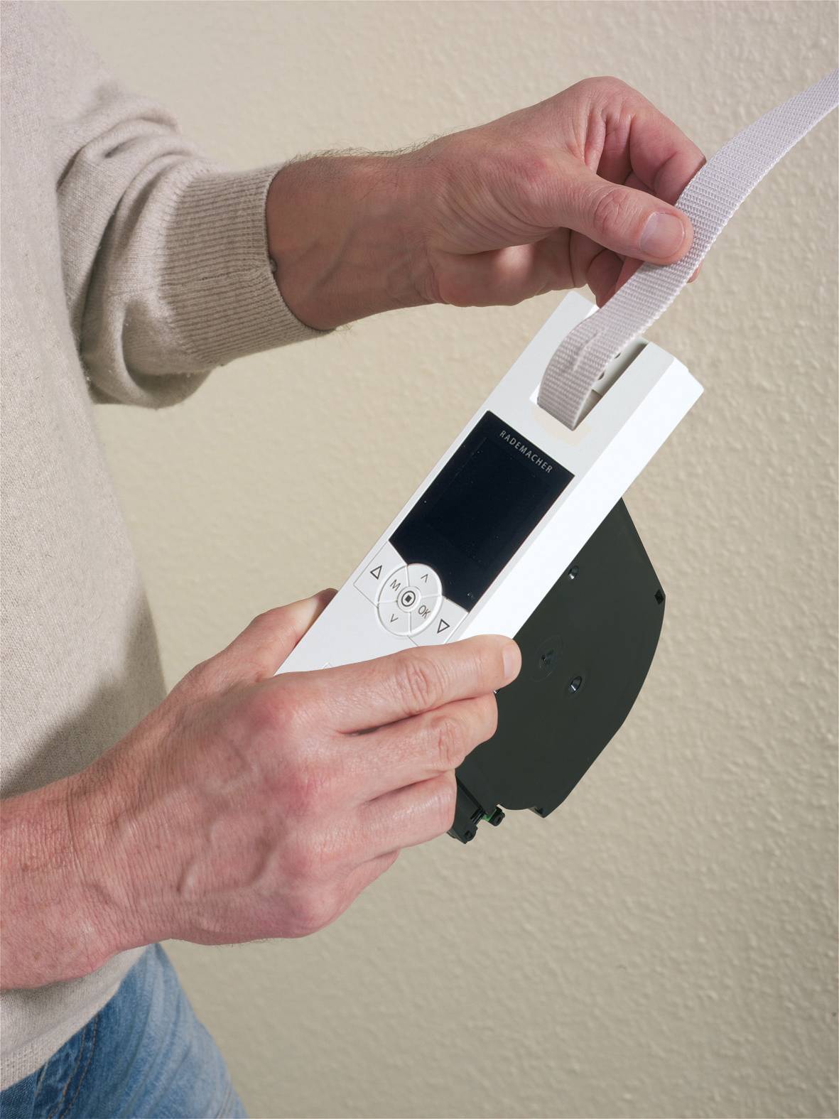 'Osoba trzyma pilot i ciągnie za pasek. Pilot posiada przyciski do sterowania. Tłem jest jasna ściana.'