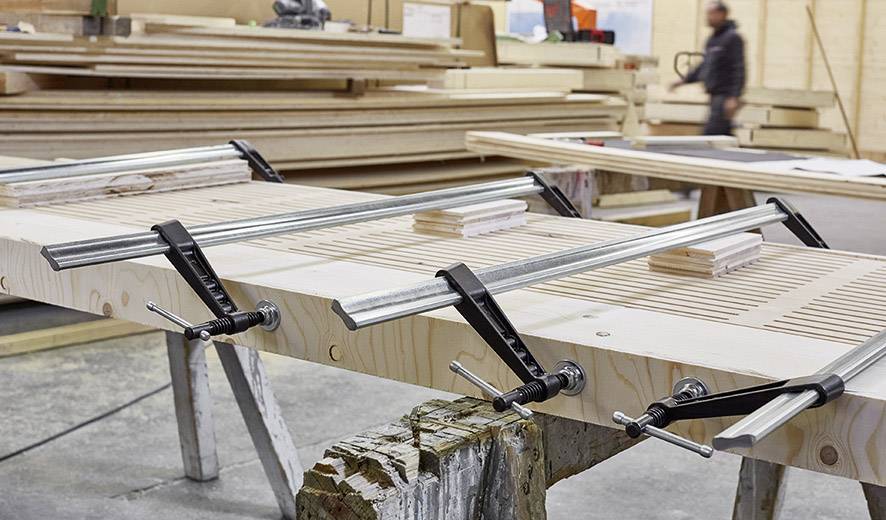 Drewniana płyta jest obrabiana w warsztacie za pomocą metalowych zacisków. W tle stoją ułożone w stos deski, a w nieostrej perspektywie widać osobę.
