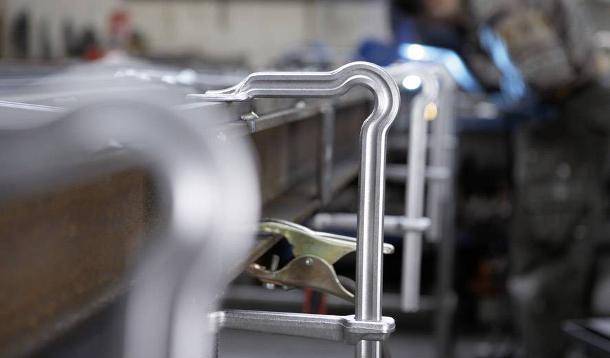 Metalowe zaciski przytrzymują elementy obrabiane na stole w warsztacie. W tle niewyraźna postać pracuje nad projektem.