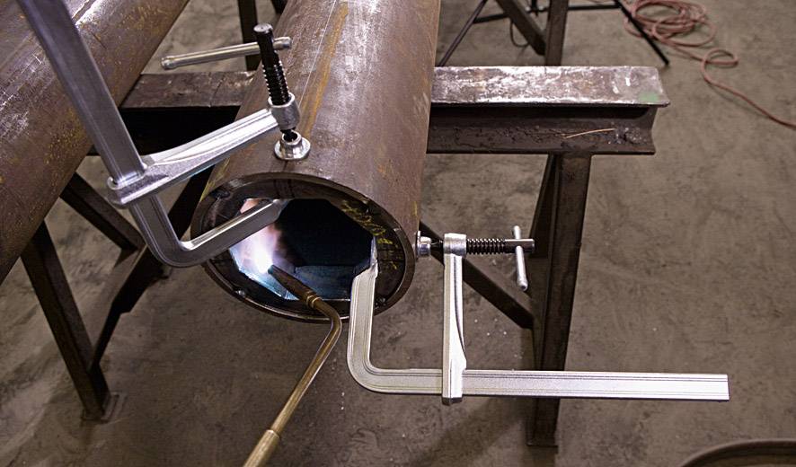 Duża rura jest zamocowana zaciskiem. Do spawania używa się płomienia. W tle widoczne są narzędzia.
