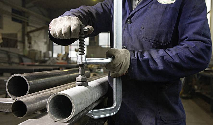 Osoba ubrana w niebieskie ubranie robocze obrabia metalowe rury za pomocą imadła w środowisku warsztatu.