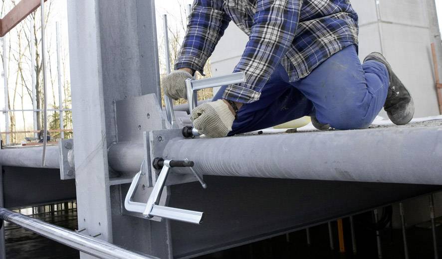 Osoba w stroju roboczym klęczy na stalowej belce i mocuje klamry.