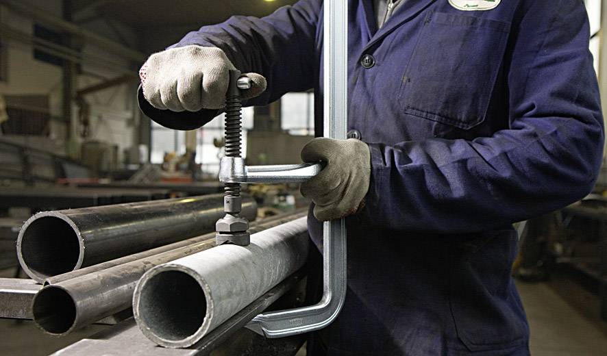 Osoba mocuje metalową rurę w warsztacie za pomocą zacisku. Ma na sobie ubranie robocze i rękawice.
