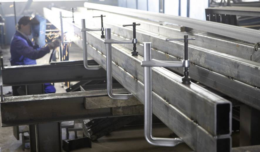 Warsztat obróbki metalu: Kilka metalowych elementów jest zamocowanych zaciskami. W tle pracuje osoba w masce ochronnej.