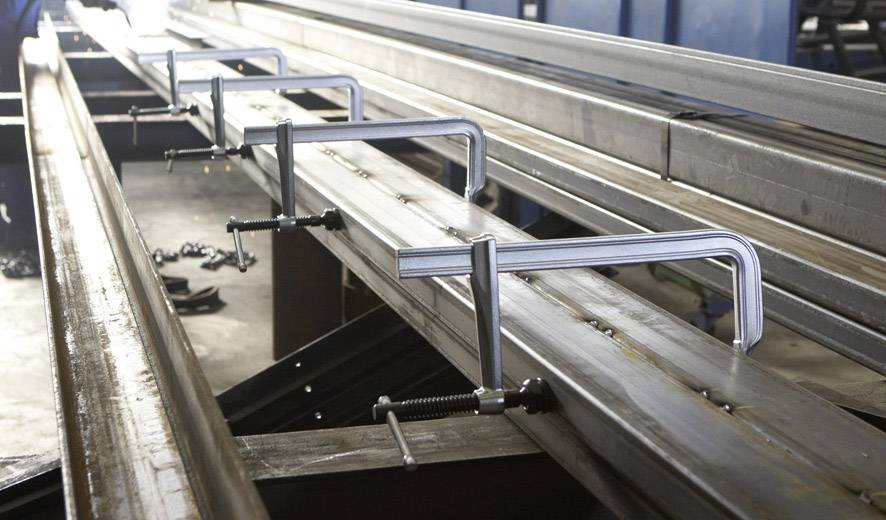 Obróbka metalu: Cztery zaciski C mocują stalowy element nośny na szynie w środowisku warsztatu.