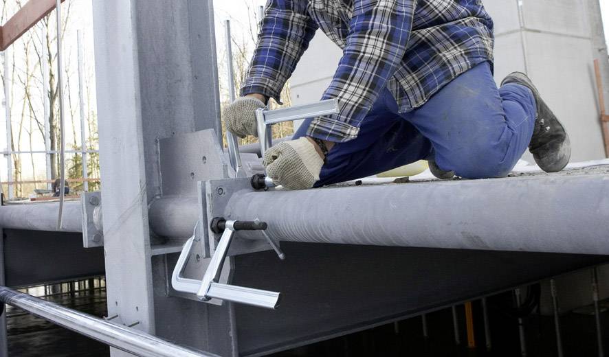 Osoba ubrana w niebieskie ubranie robocze klęczy na stalowej belce i mocuje kątownik metalowy przy pomocy ścisków.