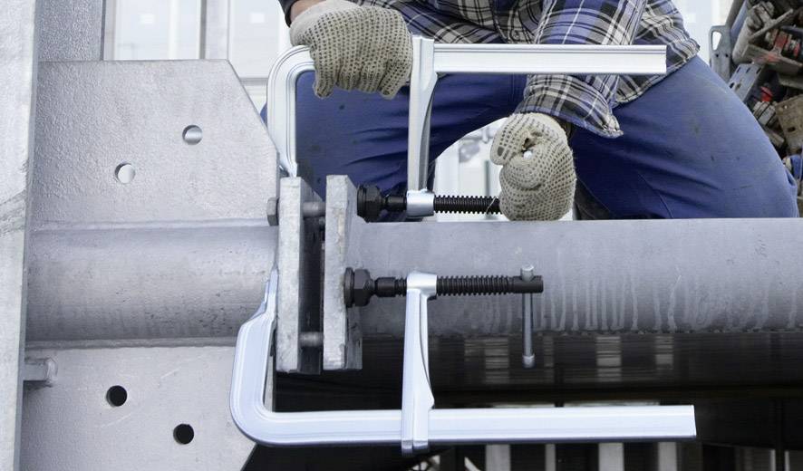 Osoba ubrana w strój roboczy i rękawice mocuje metalowe elementy do rury za pomocą dwóch ściskaczy C.