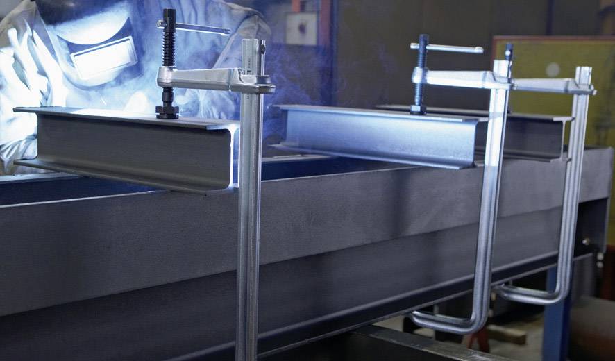 Spawacz pracuje w warsztacie, gdzie nośniki metalowe są zamocowane zaciskami. Widoczne są iskry i jasne światło.