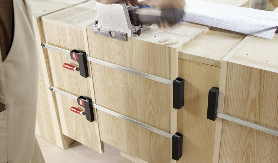 Osoba pracuje nad projektem drewnianym, używa zacisku, aby zamocować drewno na stole warsztatowym, w tle widoczne są plany.