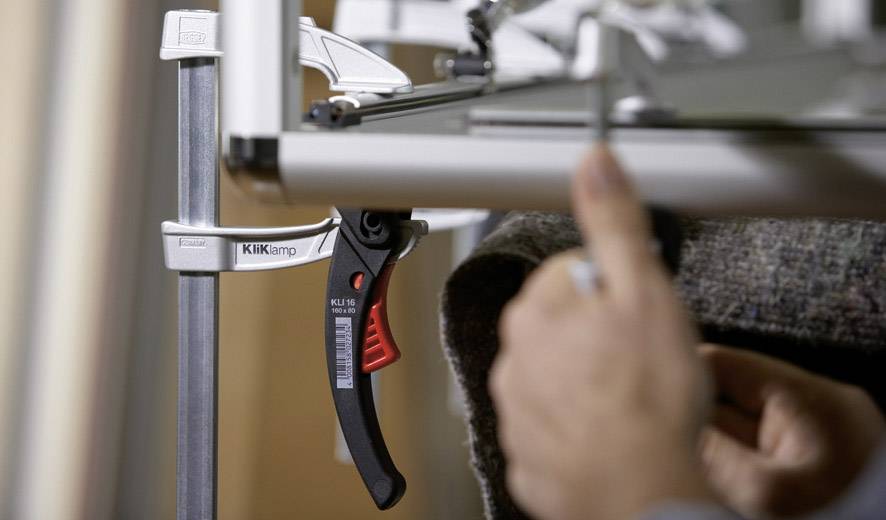 Osoba mocuje element metalowy zaciskiem do stołu. Zacisk jest czarny z czerwonym uchwytem i białym napisem.