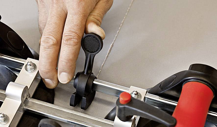 Dłoń reguluje dźwignię narzędzia, prawdopodobnie przecinarki do płytek, na metalowej szynie prowadzącej.