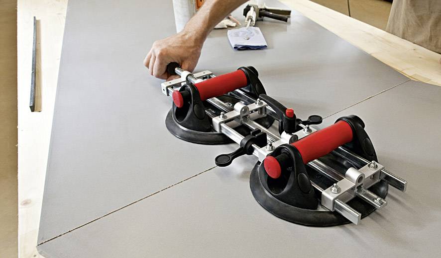 Jedna ręka używa dwóch czerwonych przyssawek, aby umieścić dużą szarą płytę na stole. Na tle widoczne są narzędzia i papier.