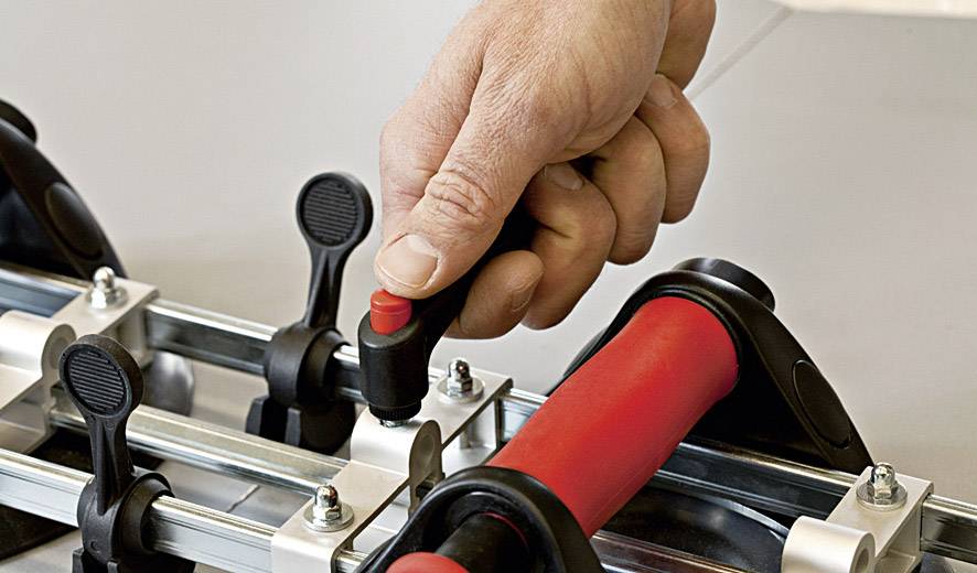 Dłoń obraca pokrętło na urządzeniu z mechanizmem dźwigniowym i metalowymi prętami, prawdopodobnie w celu regulacji lub zamocowania.