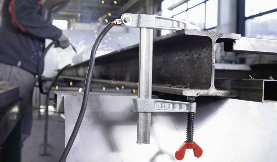 Jedna osoba spawała metal w warsztacie. Na pierwszym planie widać zacisk metalowy zabezpieczający element obrabiany.