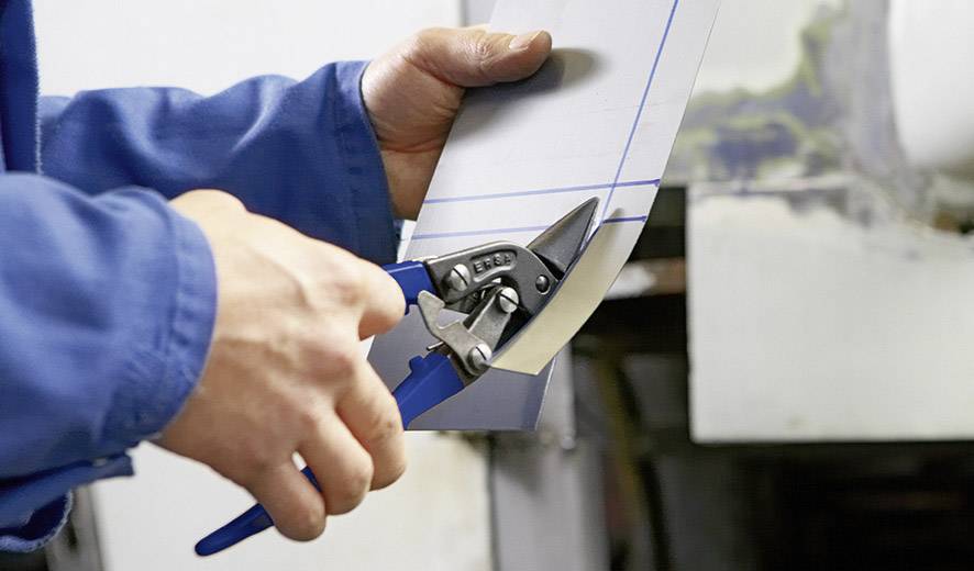 Jedna osoba przycina kawałek metalu nożycami do blachy. Ręce są w niebieskim kombinezonie roboczym.