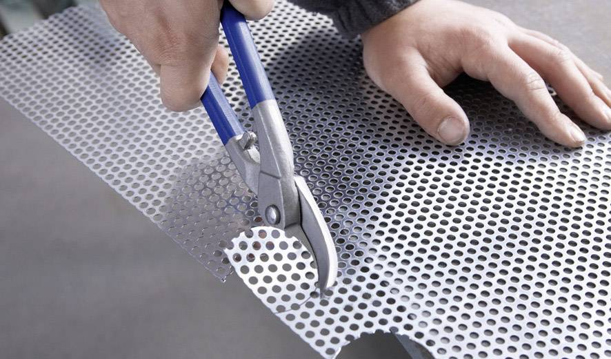 Ręka trzyma nożyce do blachy i wycina perforowaną blachę metalową. Zdjęcie przedstawia proces pracy i narzędzie w akcji.