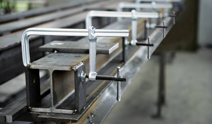 Metalowy nośnik jest mocowany za pomocą kilku zacisków śrubowych na stole warsztatowym. Zaciski utrzymują nośnik stabilnie podczas obróbki.