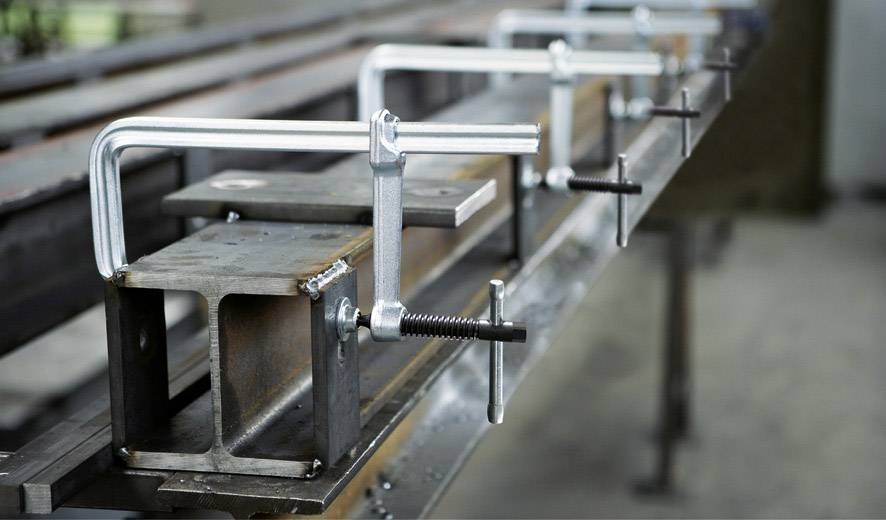 Metalowy nośnik zostaje mocno zamocowany na stole roboczym za pomocą kilku metalowych zacisków. Otoczenie ma charakter przemysłowy.