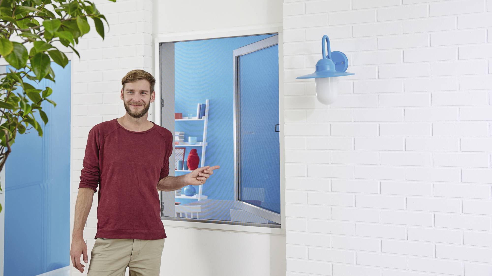 Mężczyzna w czerwonym swetrze stoi przed białą ścianą z cegły i wskazuje na okno z niebieską ramą. Po lewej stronie zielone drzewo.
