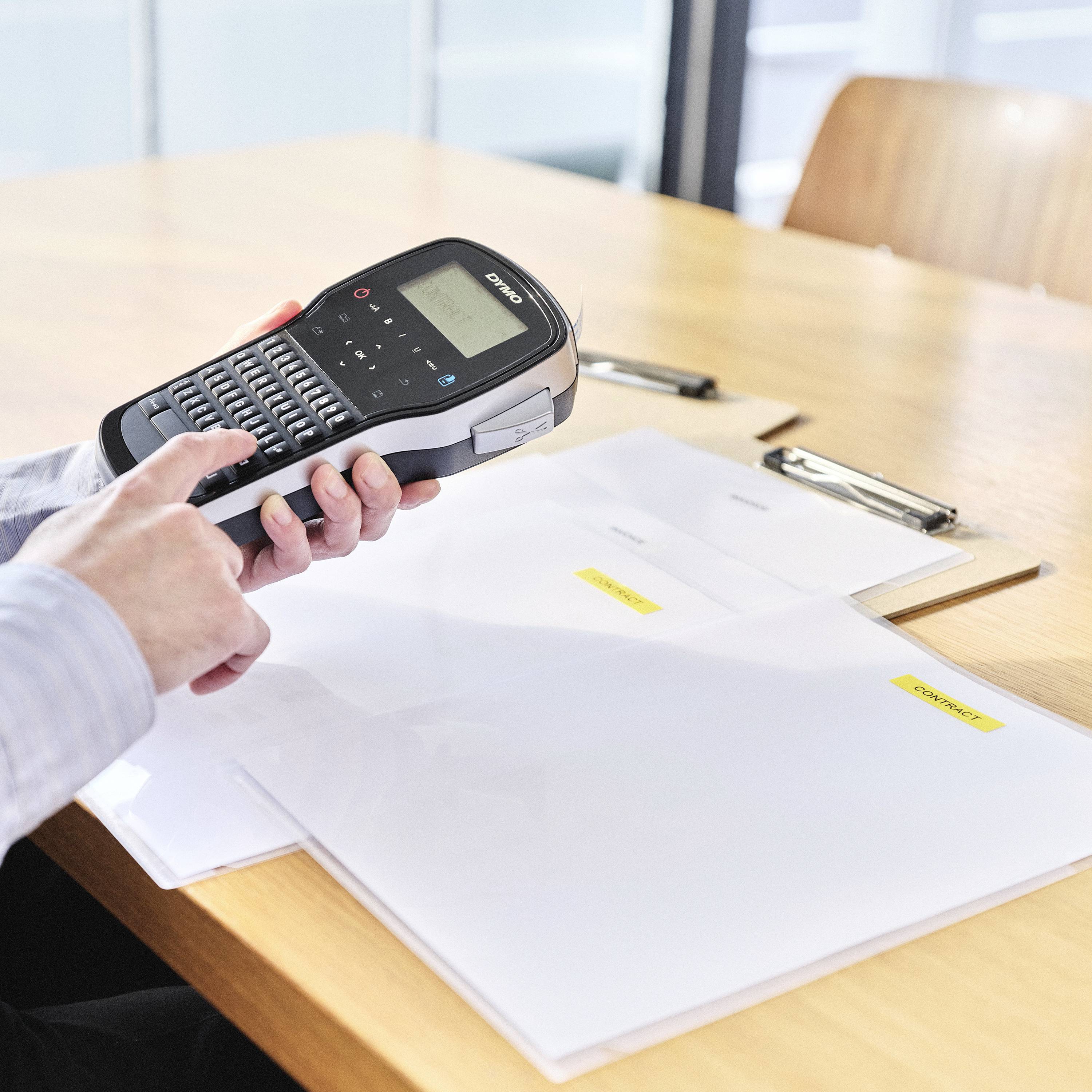 Osoba opisuje segregatory za pomocą urządzenia do etykietowania, stojąc przy stole. Widoczne są dwie teczki z dokumentami.