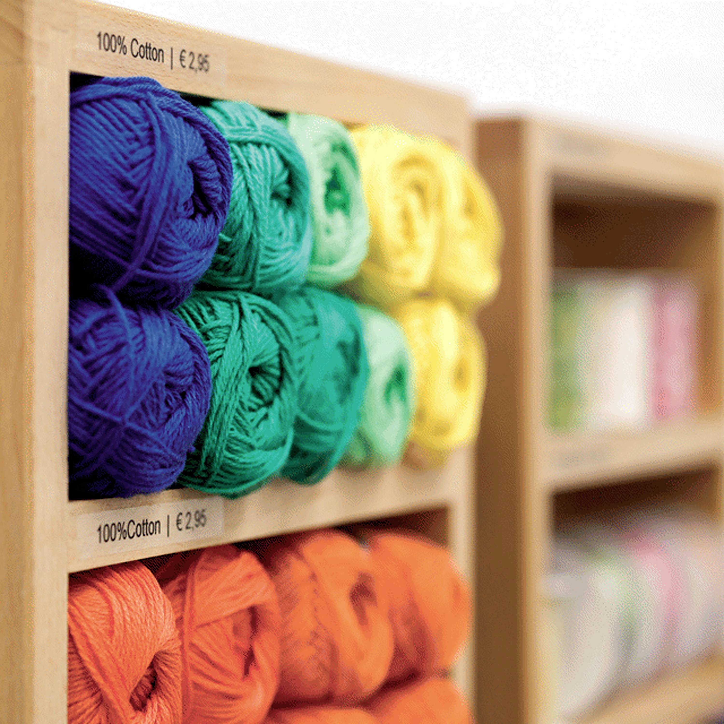 Kolorowe kłębki wełny ułożone na drewnianej półce, ułożone w gradiencie od niebieskiego do czerwonego, z cenówkami oznaczonymi '100% Cotton EUR2.95', w tle widoczne kolejne półki z wełną.